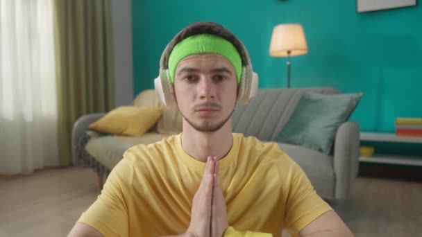 一个戴耳机的年轻人 听音乐 坐到荷花的位置 做瑜伽 一个穿着运动服的男子坐在客厅地板上的画像 — 图库视频影像