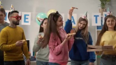 Modern ofis çalışanları pizza yiyor. Parti şapkalı ve aksesuarlı bir grup genç bir partide ofiste. Kapatın. Parti, doğum günü, ofis partisi