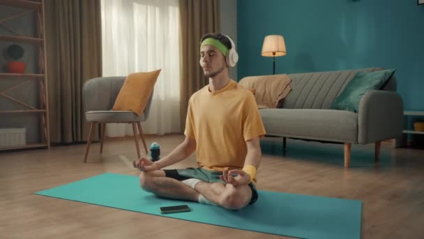一位身穿运动服的年轻人坐在客厅靠近智能手机的地板上的莲花位置上 一个男人用白色无线耳机冥想 听音乐 体育概念 — 图库视频影像