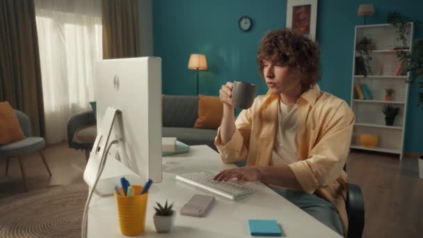 一个卷曲的小伙子坐在电脑前 喝着早晨的咖啡和茶 男人在键盘上打字 写电子邮件 上网工作 自由职业者 慢动作 — 图库视频影像