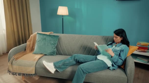 亚洲女人坐在沙发上热切地看书 一个戴着红色无线耳机的卷发小伙子正在她旁边跳舞 休息的概念在家里 慢一点 — 图库视频影像