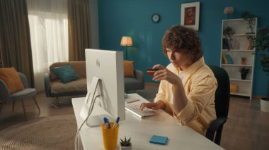 Genç bir adam online alışverişleri ödemek için bilgisayar ve kredi kartı kullanıyor. Kıvırcık saçlı adam dikkatlice veri giriyor, karttan gelen bilgileri bilgisayara aktarıyor. Evini terk etmek zorunda değilsin.