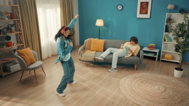 Genç kıvırcık adam kanepede kitap okuyor. Yanındaki Asyalı kadın güçlü bir şekilde kırmızı kablosuz kulaklık takarak dans ediyor. Evde dinlenme, rahatlama konsepti.