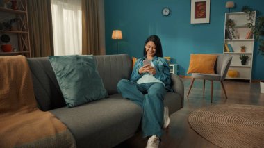 Genç bir Asyalı kadın elinde telefonla kanepede oturuyor. Bir kadın gülümseyerek mesaj yazıyor, sohbet ediyor, sosyal ağları tarıyor, görüntülere, videolara bakıyor.