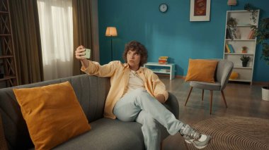 Genç bir adam akıllı telefondan selfie çekiyor. Kıvırcık saçlı bir adam oturma odasındaki kanepede oturuyor. Elinde telefon var.