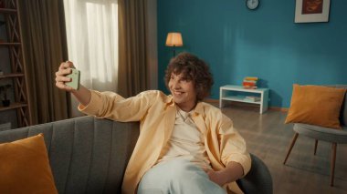 Genç, gülümseyen bir adam selfie çeker, akıllı telefondan video çeker. Kıvırcık saçlı bir adam oturma odasındaki kanepede oturuyor. Elinde telefon var.