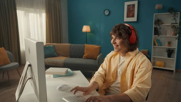 一个穿着红色无线耳机的心满意足的年轻人坐在电脑前 一个年轻人玩网络游戏 看视频 听音乐 靠近点 — 图库照片