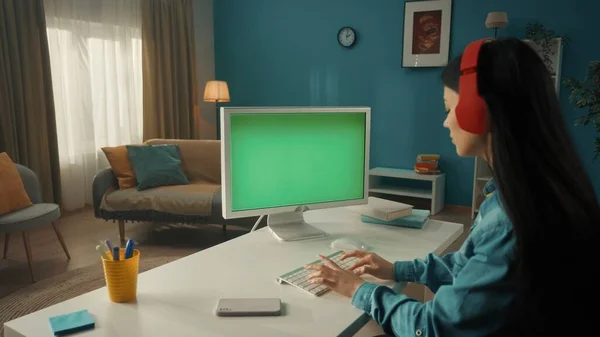 一位年轻的亚洲妇女在电脑上工作 电脑上有一个绿色的屏风 一个留长发的黑发女人坐在一张红色无线耳机的桌子旁 侧视图 通信的概念 — 图库照片