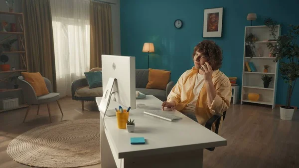 一个卷曲的小伙子坐在电脑前 耳朵上拿着智能手机 一个人一边在电脑上工作一边在讲电话 连接的概念 — 图库照片