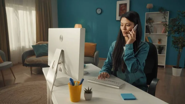 一位长相严肃的年轻的亚洲女人坐在电脑前 耳朵上戴着智能手机 一个女人在电脑前工作的时候在讲电话 通信的概念 — 图库照片