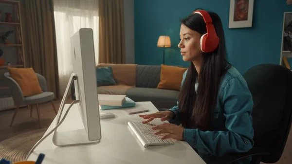 一个身穿红色无线耳机的亚洲女人在电脑上工作的侧视图 一个年轻的女人玩网络游戏 看视频 听音乐 靠近点 — 图库照片