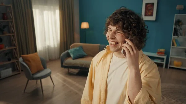 一个心满意足的年轻人用智能手机说话 笑着说 客厅里一个带着电话的卷发男子的画像 — 图库照片