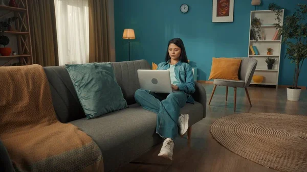 魅力的なアジアの女性は彼女の膝のラップトップでリビングルームのソファに座っています 女性の作品は インターネットを閲覧し ビデオを見ています リモートワークフリーランサー — ストック写真