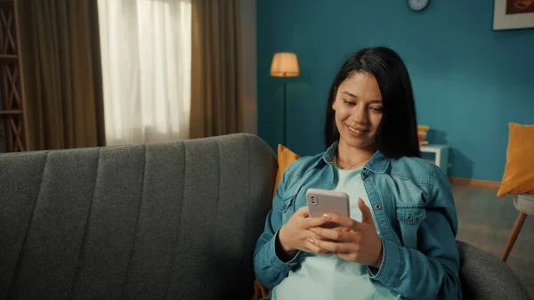 一个年轻的亚洲女人坐在沙发上 手里拿着电话 一个女人在输入一个信息 浏览社交网络 滚动通过饲料 生活中一个微笑的女人的画像 — 图库照片