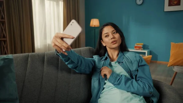 魅力的なアジアの女性は スマートフォン ポーズで自撮りを取ります 若い女性がリビングルームでソファに座ってる携帯を手に — ストック写真