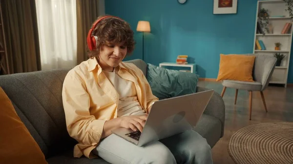 若い男が膝の上にノートパソコンと赤い無線ヘッドフォンを着てソファに座っている テキストを入力している メッセージ チャット 音楽を聞いて オンラインゲームをプレイ 閉じろ — ストック写真