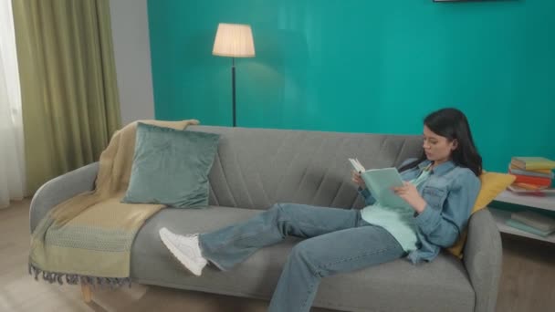 亚洲女人坐在沙发上热切地看书 一个戴着红色无线耳机的卷发小伙子正在她旁边跳舞 休息的概念在家里 慢一点 — 图库视频影像