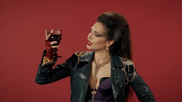 フリークの女性は手にワインのグラスを持ち 冷静にそれを見て唇をなめる 明るいメイク ビュッフェ 赤のスタジオでチェーン付きの革のジャケットを持つ女性の肖像画 — ストック動画