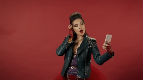 非公式な女性は自撮りをし 赤い背景のスタジオでスマートフォンでビデオを撮影します 女性はポーズをとり 髪をまっすぐにし 唇を出してカメラの前にキスを吹く ビデオ2 — ストック動画
