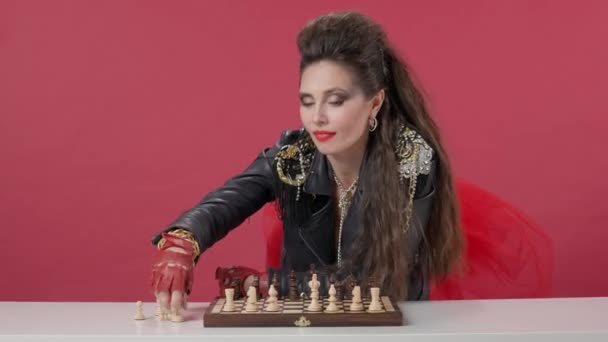 女性はチェッカーボードにチェスの駒を置き テーブルから立ち上がり すべてがゲームの準備ができていることを彼女の手で示しています スタジオでチェスをしたテーブルの上の赤い背景の女性 — ストック動画