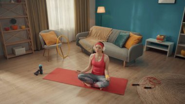 Genç bir kadın Lotus pozisyonunda gözleri kapalı bir şekilde oturur. Pembe eşofman giyen, saç bandı takan ve kablosuz kulaklık takan kızıl saçlı bir kadın evde yoga yapıyor. Yukarıdan bak. Spor.