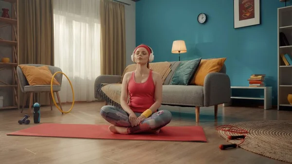 一位年轻貌美的女子盘腿坐在荷花的位置上 闭着眼睛 一位身穿粉色运动服 头带和白色无线耳机的红头发女人在家里练习瑜伽 — 图库照片