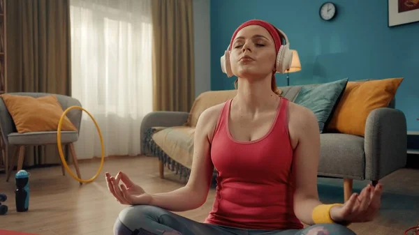 一位身穿粉色运动服 头带和白色无线耳机的红头发女人在家里练习瑜伽 年轻妇女盘腿坐着 闭上眼睛 莲花的位置体育概念 — 图库照片
