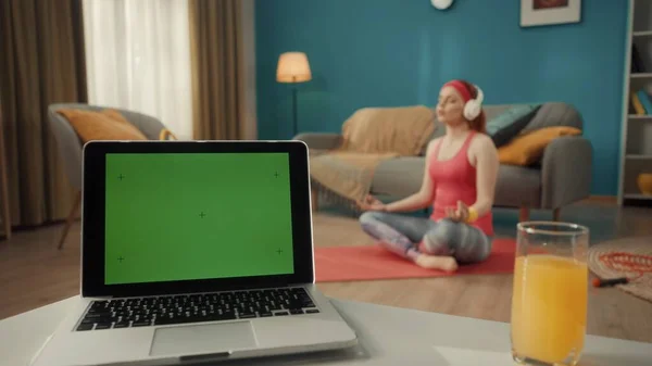 客厅的桌上放着一台装有绿色屏幕和一杯果汁的笔记本电脑 在背景音乐中 一个戴耳机的女人坐在莲花的位置上 听网上一课 体育概念 — 图库照片