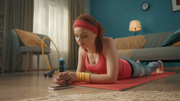 一位穿着粉色运动服和运动帽带的年轻女子躺在地板上的运动垫上 一个靠在手肘上的年轻女人看着电话 靠近点体育视频课程 在线体育 — 图库照片