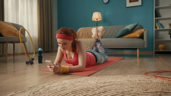 一位穿着粉色运动服的红头发年轻女子躺在客厅的地板上 一个女人手里拿着一个电话 看着照片 填写一个电子训练日志 — 图库照片