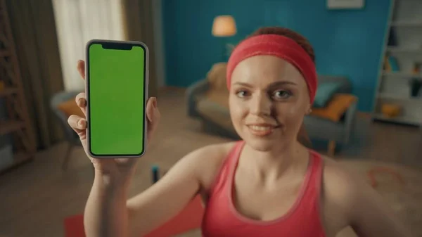 一个红头发女人的画像 穿着粉红色的运动服 头带在客厅里做完运动后的特写 一个女人用绿色屏幕显示智能手机的屏幕 广告领域 工作空间 — 图库照片