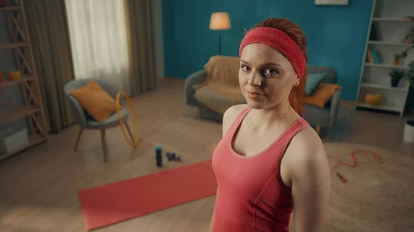 一个红头发的女人穿着粉红色的运动服 戴着运动帽带 躺在客厅里 地板上有运动器材 一个苗条迷人的女人直视前方 靠近点 — 图库照片