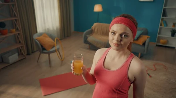 一个红头发女人穿着粉色运动服的画像 客厅里戴着运动帽带 地板上有运动器材 一个纤细的女人手里拿着一杯果汁 直视前方 — 图库照片