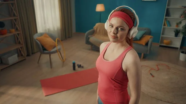 客厅里一个头戴白色无线耳机的红头发女人的画像 地板上有运动器材 一个穿着粉色运动服的年轻女子直视前方 靠近点 — 图库照片