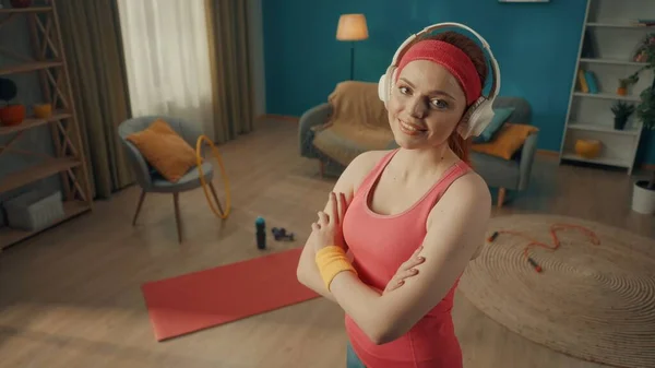 客厅里一个头戴白色无线耳机的红头发女人的画像 地板上有运动器材 一位身穿粉色运动服的年轻女子 双臂交叉在胸前 直视前方 — 图库照片
