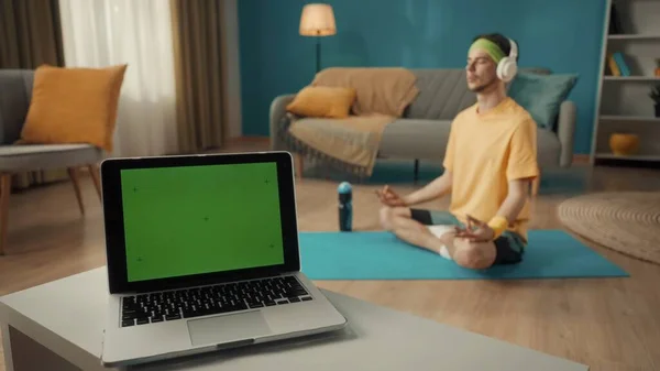客厅的桌上有绿色屏风的笔记本电脑 在背景中 一个穿着运动服和白色无线耳机的男人坐在莲花的位置上 体育概念 瑜伽课 广告领域 — 图库照片