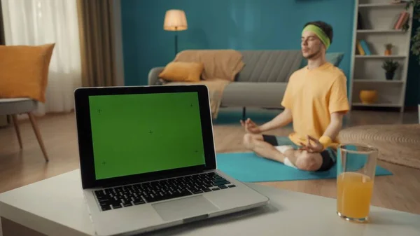客厅的笔记本电脑上有绿色屏风 桌上有一杯果汁 在背景中 一个身穿运动服 头戴运动头巾的男人坐在一个莲花的位置上 体育概念 瑜伽课 — 图库照片