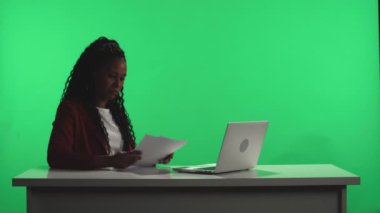 TV haber programı. Siyah bir stüdyoda, yeşil bir ekrana karşı bir Afro-Amerikan TV sunucusu bir dizüstü bilgisayar ve kağıt üzerinde bir program planı kullanarak yayına hazırlanıyor. Işık yanıyor ve kadın...