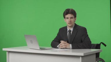 TV Canlı Haber Programı: Erkek Sunucu Muhabiri. Tekerlekli sandalyedeki spiker konuşuyor, dinliyor, yeşil ekranın arka planına kafa sallıyor. Haber Odası Stüdyo Konsepti. Reklam alanı, çalışma alanı maketi