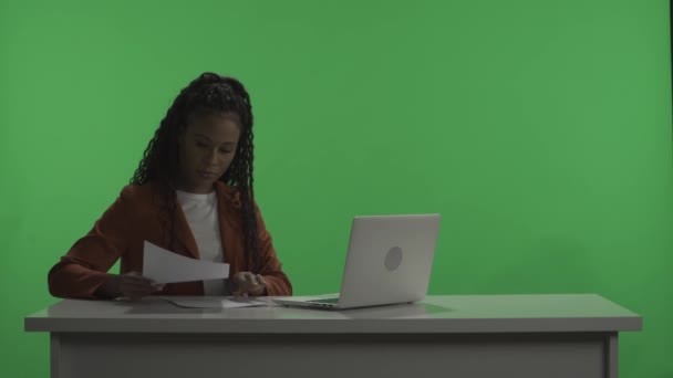 电视节目主持人坐在黑暗的演播室里的一张桌子旁 面对着一个绿色的屏幕 一名妇女为广播做准备 修改节目计划 在笔记本电脑上打字 用纸张输入信息 广告领域 — 图库视频影像