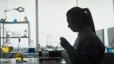 Bilimsel bir deney yapan kadın bir bilim adamının karanlık silüeti. Pipet kullanan bir kadın cam şişeye temiz bir sıvıyla yeşil bir reaktör ekler. Kadınların eli matarayı sıkıyor.