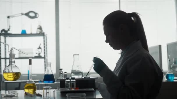 一个正在进行科学实验的女科学家的黑暗轮廓 一个女人使用管道将添加一个绿色试剂到玻璃瓶与清澈的液体 女人的手在靠近时握着一个瓶子 — 图库视频影像