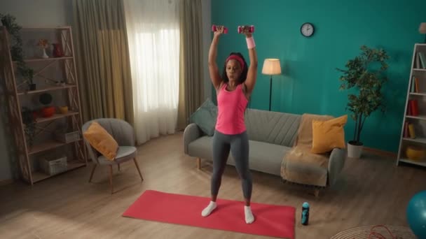 一位身穿运动服的非洲裔美国妇女抬起胳膊 头顶上方的哑铃穿过两边 年轻女子站在客厅的运动垫上锻炼肩膀肌肉 — 图库视频影像