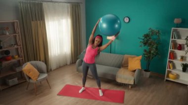 Afrikalı bir Amerikalı kadın düz kollarıyla bir fitness topu kaldırıyor ve yan eğiliyor. Genç bir kadın spor için evine gidiyor. Spor konsepti, ev sporu. Yukarıdan görüntüle
