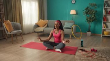 Genç bir Afrikalı Amerikalı kadın lotus pozisyonunda oturur gözleri kapalı ve yavaşça nefes alır ve nefes verir. Atletik kadın oturma odasında yerde bir spor minderinin üzerinde oturuyor. Spor konsepti. Yoga