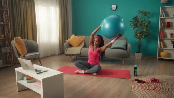 一个年轻的女人盘腿坐在一个运动垫上 在笔记本电脑上看视频课 非洲裔美国妇女抬起直胳膊 头顶上方有一个健身球 并做侧弯 家庭健康 — 图库视频影像