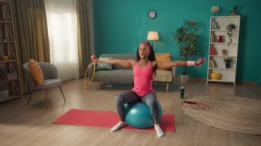 Afrika kökenli Amerikalı bir kadın spor salonunda oturuyor ve evde dambıllarıyla egzersiz yapıyor. Spor topunun üzerinde oturan bir kadın, kollarının ve omuzlarının kaslarını çalıştırır. Spor konsepti, ev fitness