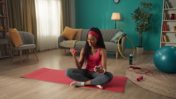 アフリカ系アメリカ人の女性がリビングの床の上のスポーツマットに足を組んで座っている 女性は片手にダンベルを持ち 手を動かすとスマートフォンを持っている — ストック動画