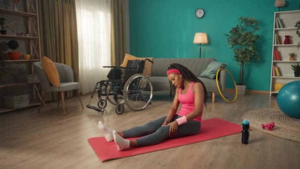 一位非洲裔美国妇女坐在轮椅旁边客厅的地板上 一个年轻的女人给自己做足底按摩 身体康复的概念 — 图库视频影像