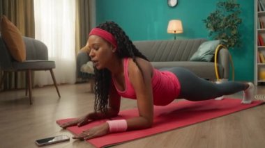 Spor giyimli Afro-Amerikalı kadın telefonun önünde yerde yatıyor. Bir kadın telefonunda zamanlayıcı başlatır ve dirseklerinin üzerine tahta bir konuma gelir. Ev fitness konsepti, online spor